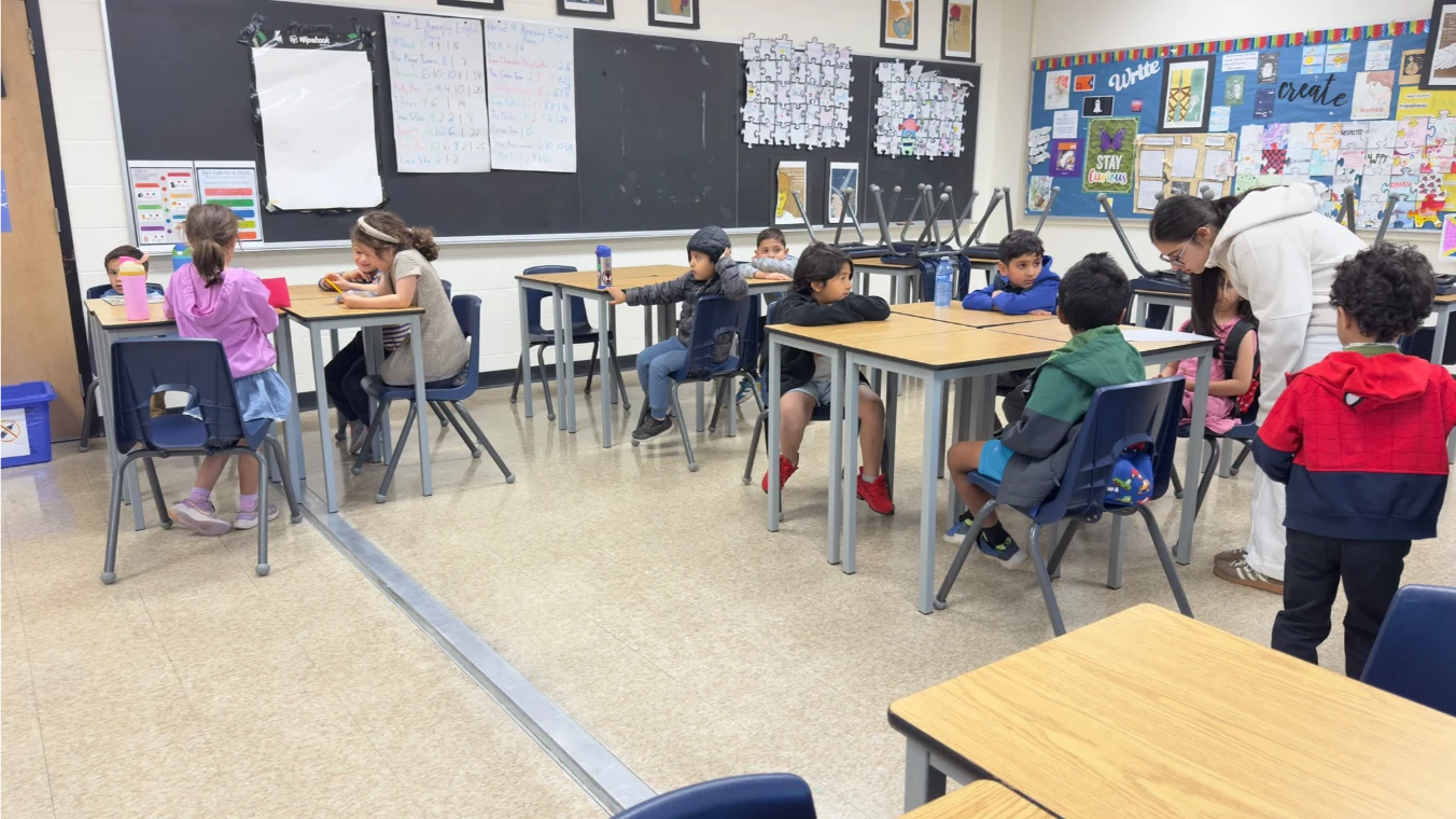 An instructor assisting a child at a desk while students participate in a small-group classroom session.